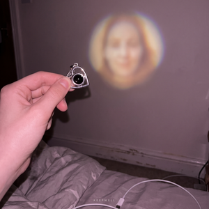 The pendant held up in one hand in a darkened bedroom, with the picture from its lens stone cast on the wall behind — a soft, warm circle with a face inside it.